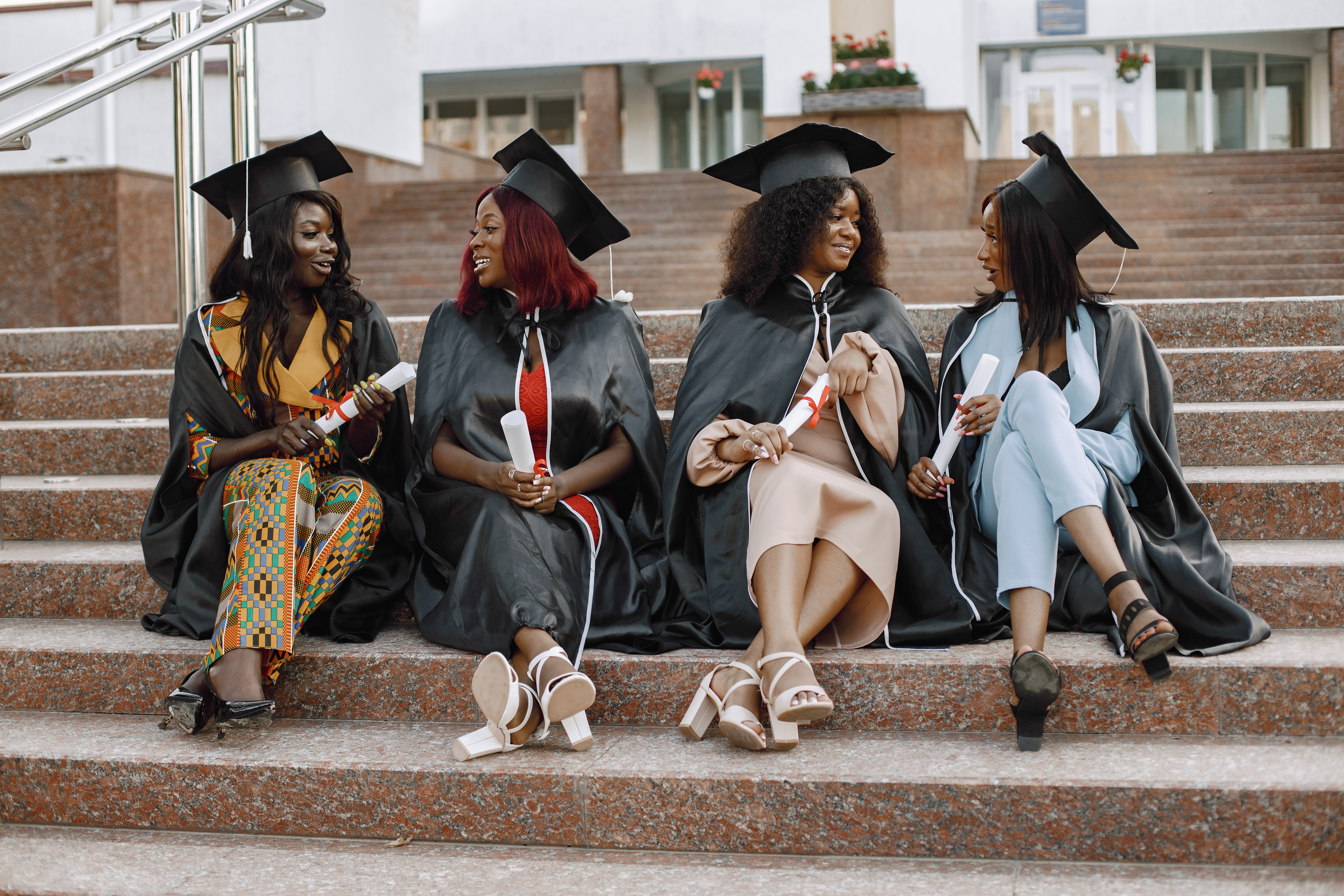 Group of young afro american female students dressed in black graduation gown. Campus as a background. Girls sitting on a stairs and talking.