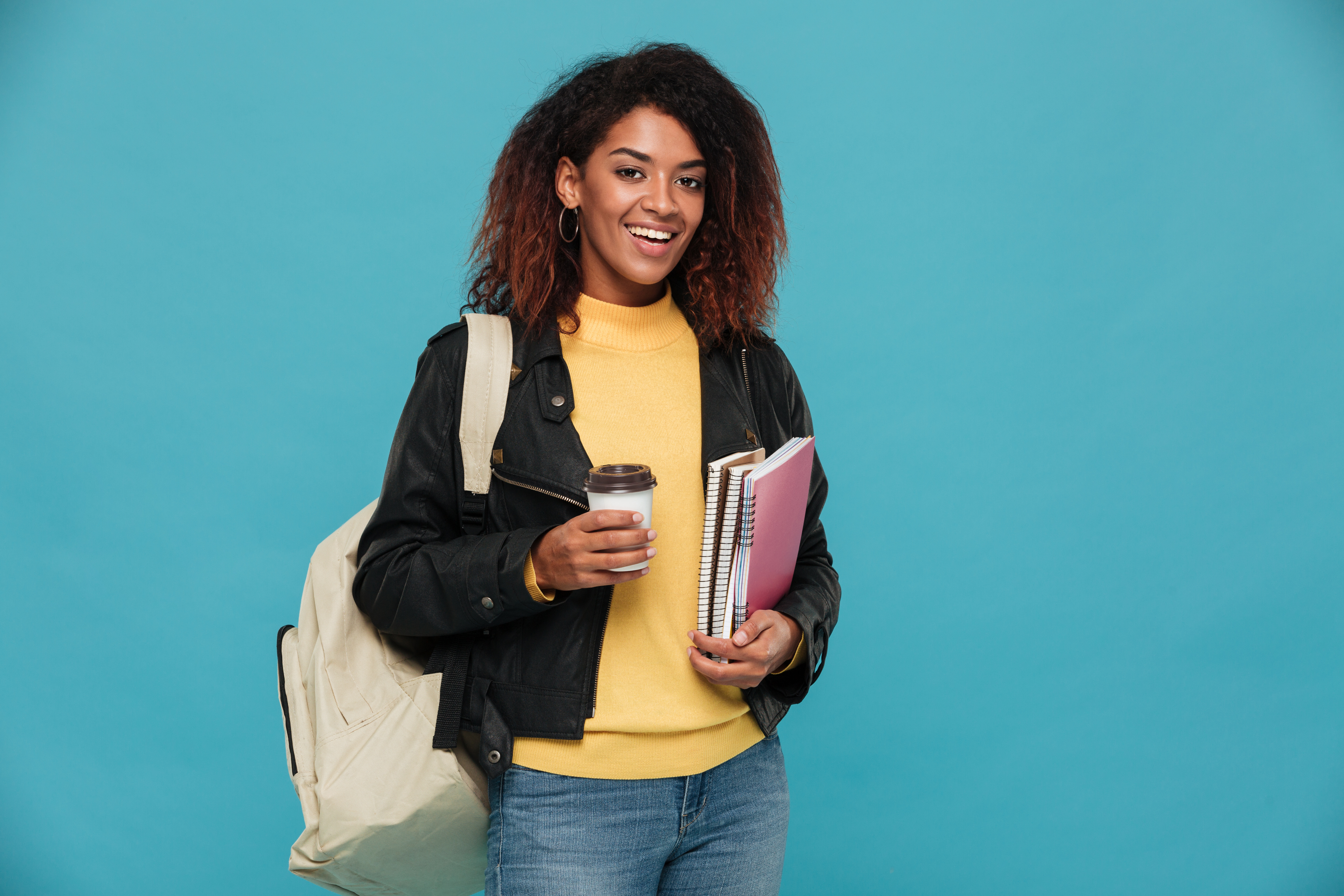 Photo of happy beautiful young african woman student standing isolated over blue background. Looking camera holding notebooks and coffee.